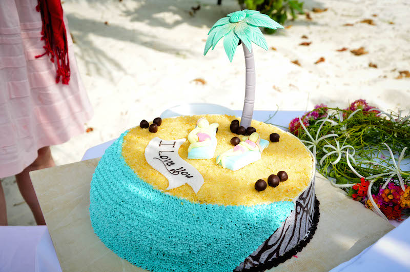 The cake is decorated with a sandy beach scene. It includes a palm tree, coconuts, two figurines and a banner reading 'I Love You'